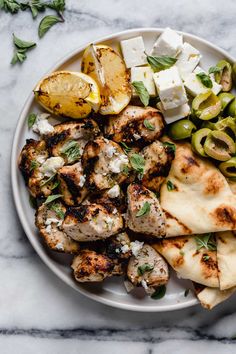 a white plate topped with chicken, potatoes and olives next to feta cheese