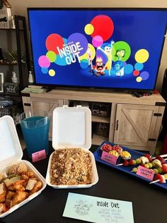 a table topped with containers filled with food next to a flat screen tv on top of a wooden entertainment center