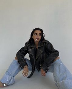 a woman sitting on the ground with her legs crossed wearing jeans and a leather jacket