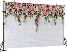 a white brick wall with pink and yellow flowers hanging from it's sides next to a black metal tripod