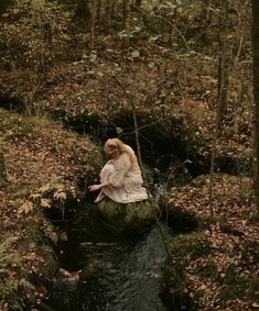 a woman sitting on top of a rock in the middle of a river surrounded by trees