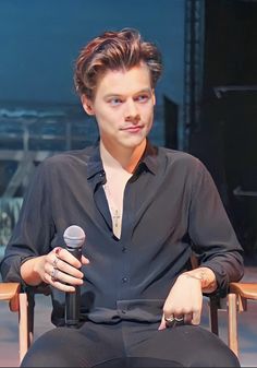 a man sitting in a chair holding a microphone and looking at the camera while wearing a black shirt