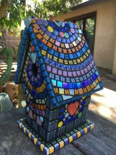 a birdhouse made out of colorful tiles on a wooden table in front of a house