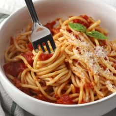 a white bowl filled with spaghetti and sauce on top of a table cloth next to a fork