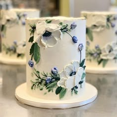 three white wedding cakes decorated with blue flowers and greenery on a silver countertop