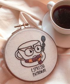 a cup of coffee sitting next to a cross stitch embroidery on a white cloth with the words espresso patronnoi