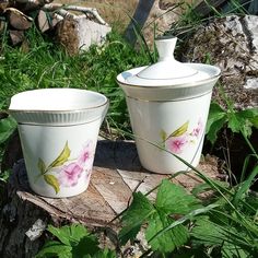 two white ceramic containers sitting on top of a tree stump in the grass with pink flowers painted on them