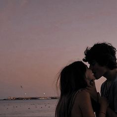 two people standing next to each other in front of the ocean at dusk with birds flying overhead