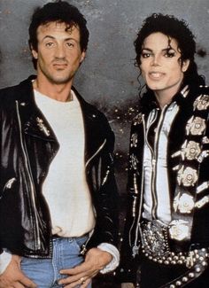 two people standing next to each other in front of a cement wall and wearing black leather jackets