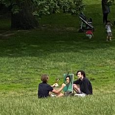 two people sitting in the grass with an easel on their lap and one person painting