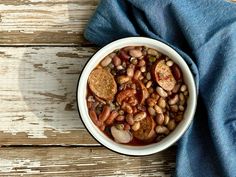 a white bowl filled with beans and meat on top of a blue cloth next to a wooden table