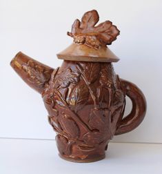 a carved teapot with a wooden lid and leaves on the top, sitting against a white wall