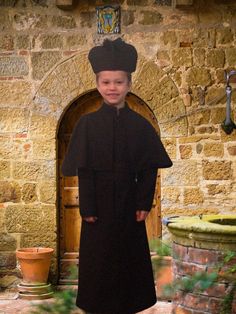 a young boy in a black robe and hat standing next to a stone building with an open door