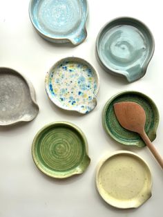 six different colored dishes with wooden spoons in them on a white surface, one is empty and the other has blue speckles