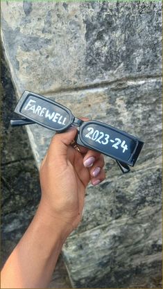 a person holding up two glasses with the word farewell written on them in front of a stone wall