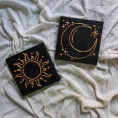 two black and gold square coasters on a white sheet with the sun and moon painted on them