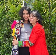 two women standing next to each other in front of some bushes and green plants with their arms around each other