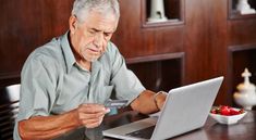 an older man sitting at a table with a laptop and credit card in front of him