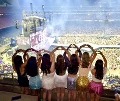 four girls standing in front of a crowd at a concert holding their hands up to the sky