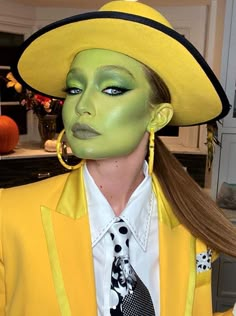 a woman in a yellow suit and hat with green paint on her face, wearing large earrings