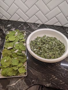 a bowl of spinach next to a tray of leaves on a marble counter top