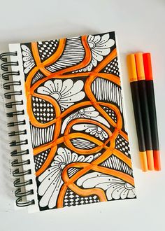 an orange and black spiral notebook next to two markers on a white surface with a design in the middle