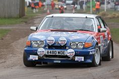 a blue car driving down a dirt road next to other cars and people in orange vests