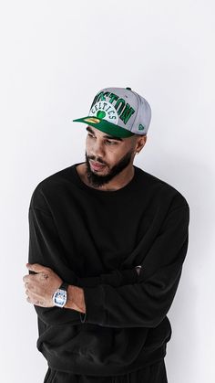 a man with his arms crossed wearing a black sweatshirt and green hat, standing in front of a white wall