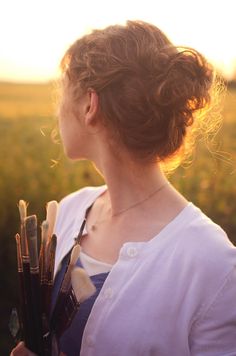 a woman holding paint brushes in her hands and looking at the sun setting behind her