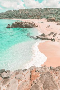 people are walking on the beach next to the clear blue water and rocky shore line