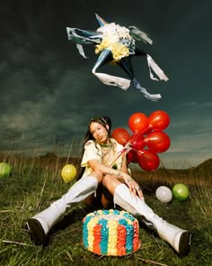 a woman sitting in the grass with balloons and a cake