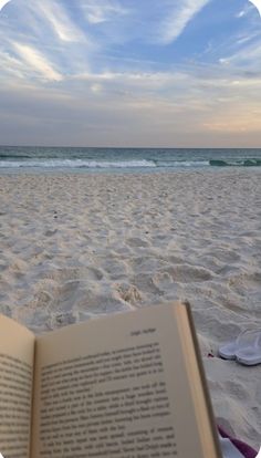 an open book sitting on top of a sandy beach next to the ocean with flip flops