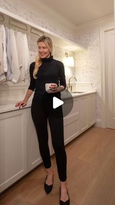 a woman standing in a kitchen holding a cup and looking at the camera while wearing black