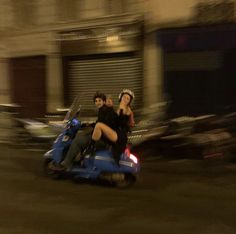 two people riding on the back of a blue scooter in front of a building