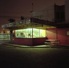 an empty street corner at night with a lit up store front and bench in the foreground