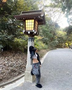 a woman standing next to a lamp post in the middle of a road with trees behind her