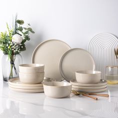 a white table topped with plates and bowls next to a vase filled with flowers on top of a counter