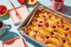 a cake in a pan with peaches and raspberries next to two cups of tea