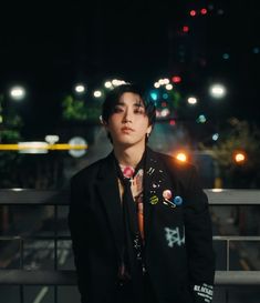 a young man in a suit and tie standing on a bridge at night with city lights behind him