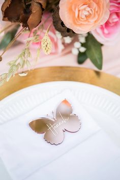 a white plate topped with a butterfly next to pink and orange flowers on top of a table