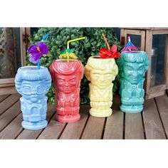 four different colored tiki cups sitting next to each other on a wooden table with plants in the background