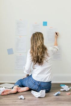 a woman sitting on the floor writing on a wall with sticky notes all over it