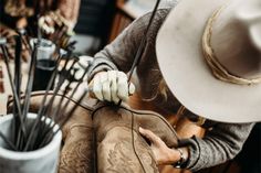 a cowboy hat is held up by his gloved hand, with other hats and utensils in the background