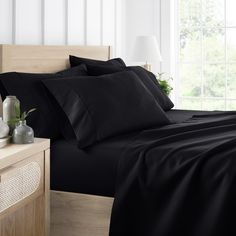 a bed with black sheets and pillows next to a night stand in front of a window