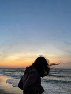 a woman standing on top of a sandy beach next to the ocean at sunset with her hair blowing in the wind