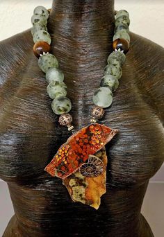 Homage to Mother Earth! Two glossed slabs of jasper (Morgan Poppy and Ocean) host an ammonite fossil as well as burnt orange crystals in this OOAK unisex statement pendant!  Flanking the pendant are two ornate copper beads.  The necklace portion consists of African recycled glass beads which are a frosted white with brown spots. Two oversized deep chocolate brown beads with ornate silver tone star bead caps serve as accents.  The back is finished with tiger's eye.  Closes with gold tone hardware Luxury Statement Necklace With Large Stone, Luxury Large Beaded Necklaces As Statement Piece, Handmade Jasper Artisan Beaded Necklaces, Handmade Jasper Beaded Necklaces Artisan Style, Handmade Jasper Beaded Necklaces, Handmade Unique Jasper Beaded Necklaces, Unique Ocean Jasper One-of-a-kind Necklace, Unique Ocean Jasper Necklace One Of A Kind, Unique One Of A Kind Ocean Jasper Necklace