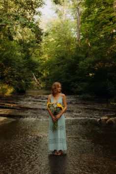 a woman standing in the middle of a river with flowers on her chest and holding a bouquet