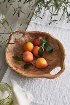 a wooden bowl filled with fruit sitting on top of a white table cloth next to a vase