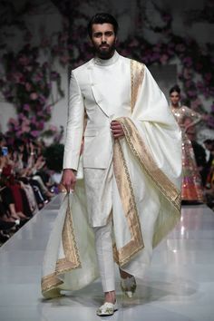 a man in a white suit and gold shawl walking down the runway