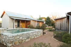 an outdoor hot tub in the middle of a garden with wooden buildings behind it and gravel path